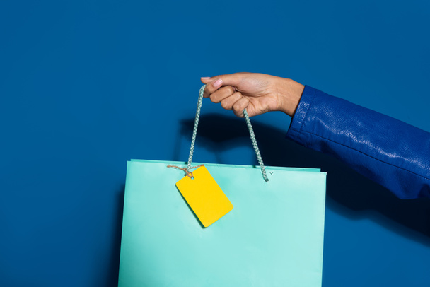 cropped view of african american woman holding shopping bag with blank label on blue - 写真・画像