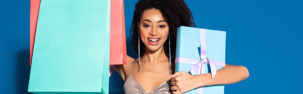 happy beautiful african american woman in silver dress holding gift box and shopping bags on blue background, panoramic shot - 写真・画像