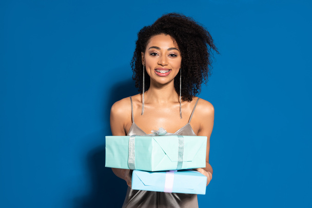 smiling beautiful african american woman in silver dress holding gifts on blue background - Photo, Image