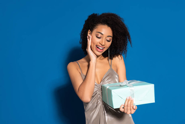 happy beautiful african american woman in silver dress holding gift on blue background - Photo, Image