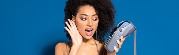 shocked beautiful african american woman in silver dress holding terminal with check on blue background, panoramic shot - Φωτογραφία, εικόνα