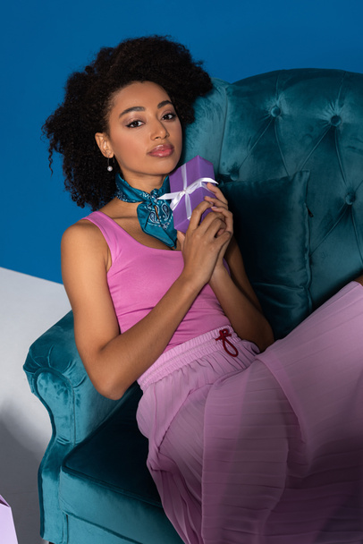 beautiful african american woman sitting in velour armchair with gift box on blue background - Fotoğraf, Görsel