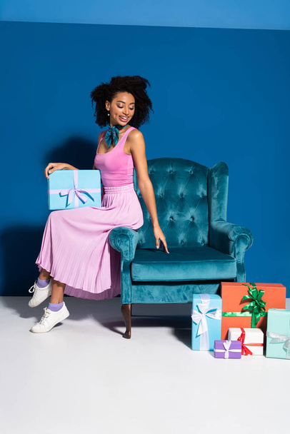 smiling african american woman sitting on velour armchair near gift boxes on blue background - Photo, Image