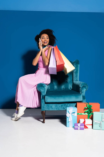 smiling african american woman sitting in velour armchair with purchases and talking on smartphone near gifts on blue background - Zdjęcie, obraz