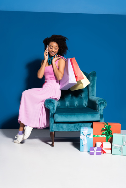 smiling african american woman sitting in velour armchair with purchases and talking on smartphone near gifts on blue background - Фото, зображення
