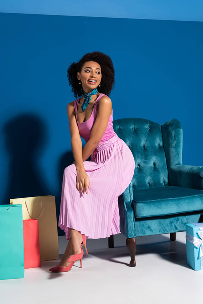 sonriente mujer afroamericana sentada en un sillón de terciopelo cerca de cajas de regalo y compras en fondo azul
 - Foto, imagen