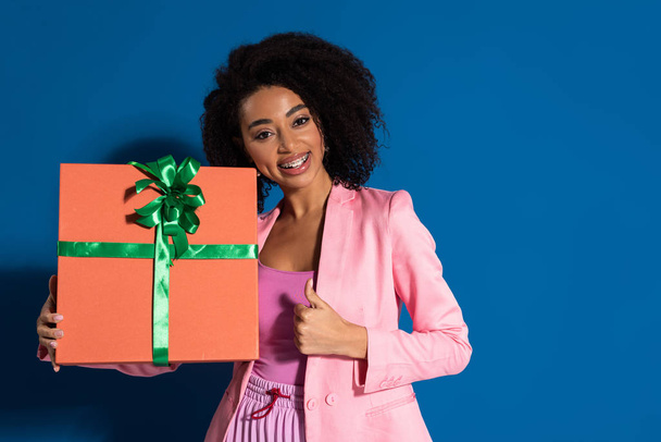 elegant smiling african american woman with gift showing thumb up on blue background - Photo, Image