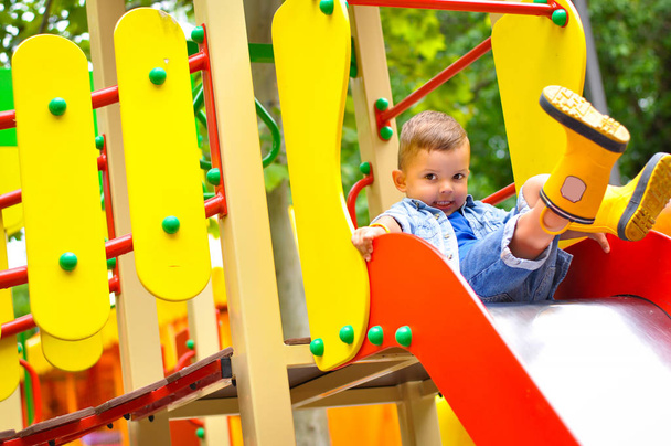 Un petit garçon mignon de trois ans joue sur le terrain de jeu et glisse sur les toboggans en bottes en caoutchouc jaune
. - Photo, image