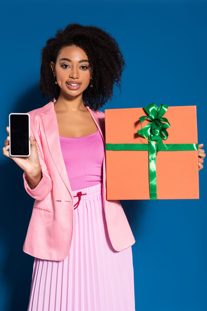 elegantní usmívající se africká americká žena s dárkem ukazující smartphone s prázdnou obrazovkou na modrém pozadí - Fotografie, Obrázek