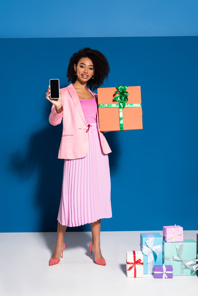 elegant smiling african american woman with gift showing smartphone with blank screen on blue background - Zdjęcie, obraz
