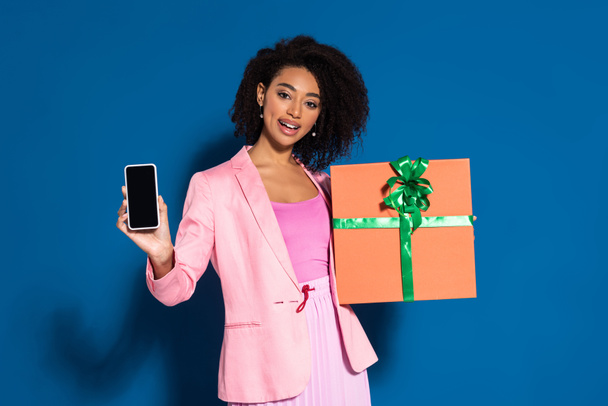 elegant smiling african american woman with gift showing smartphone with blank screen on blue background - Foto, immagini