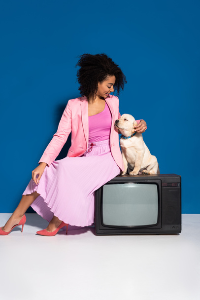 smiling african american woman sitting on vintage tv with golden retriever puppy on blue background - Фото, изображение