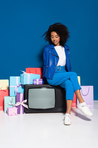 smiling african american woman sitting on vintage television near gifts and shopping bags on blue background - Foto, afbeelding