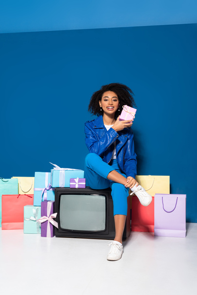 smiling african american woman sitting on vintage television near gifts and shopping bags on blue background - Φωτογραφία, εικόνα