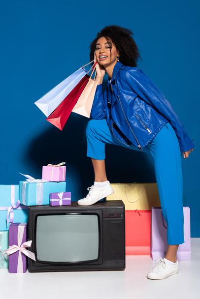 smiling african american woman sitting on vintage television with shopping bags on blue background - Foto, Bild