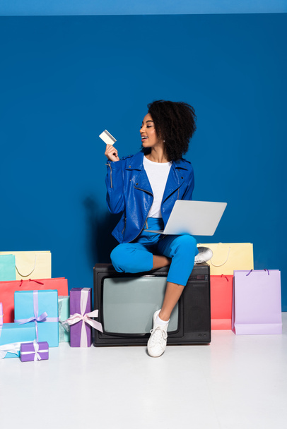sonriente mujer afroamericana sentada en la televisión vintage con computadora portátil y tarjeta de crédito cerca de regalos y bolsas de compras sobre fondo azul
 - Foto, imagen