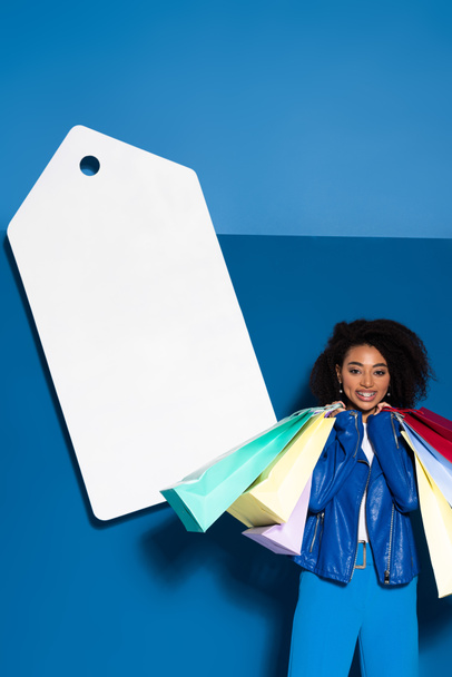 smiling african american woman with shopping bags near big blank price tag on blue background - Fotoğraf, Görsel