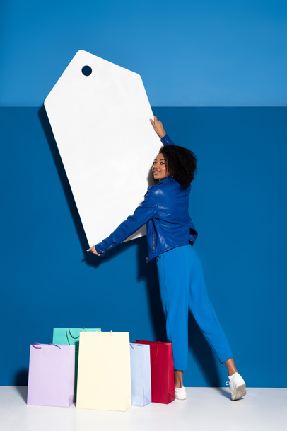 smiling african american woman holding big blank price tag near shopping bag on blue background - Foto, imagen