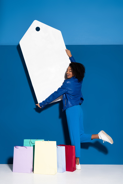 sorridente donna africana americana con in mano un grande cartellino del prezzo vuoto vicino alla borsa della spesa su sfondo blu
 - Foto, immagini