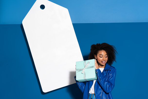 smiling african american woman with gift near big blank price tag on blue background - 写真・画像