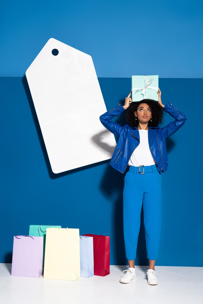 africká americká žena s dárkem v blízkosti velké prázdné cenové značky a nákupy na modrém pozadí - Fotografie, Obrázek
