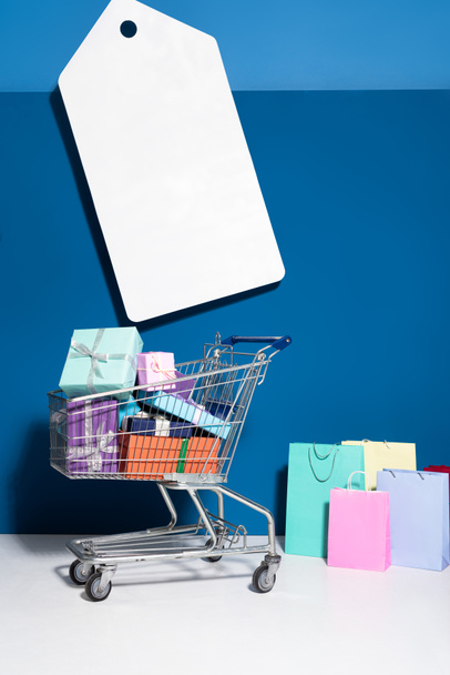 shopping bags, trolley with gifts and big blank price tag on blue background - Photo, Image