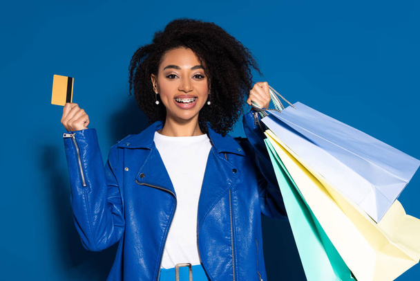 happy african american woman with credit card and shopping bags on blue background - Zdjęcie, obraz