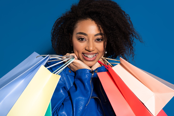 smiling african american woman with braces and shopping bags isolated on blue - 写真・画像
