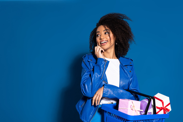 smiling african american woman holding shopping basket with gifts and talking on smartphone on blue background - Foto, Imagem