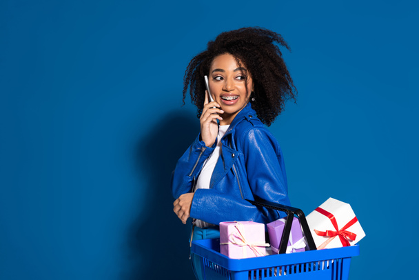 smiling african american woman holding shopping basket with gifts and talking on smartphone on blue background - Photo, Image