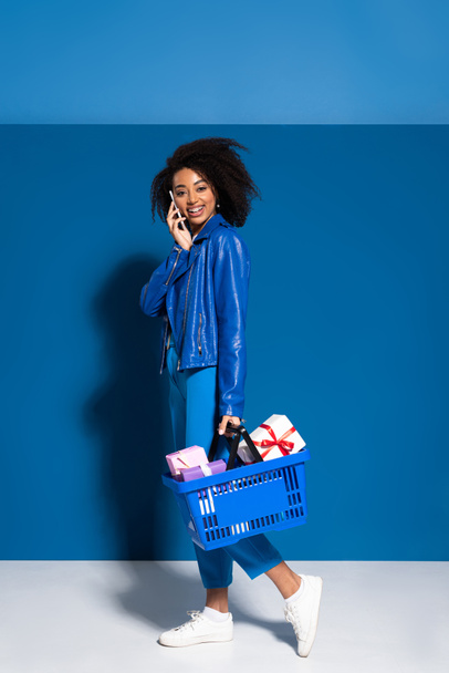 smiling african american woman holding shopping basket with gifts and talking on smartphone on blue background - Zdjęcie, obraz