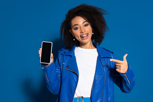 smiling african american woman pointing with finger at smartphone with blank screen on blue background - Photo, Image