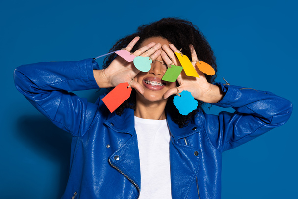 smiling african american woman covering eyes with empty labels on hands on blue background - Photo, Image