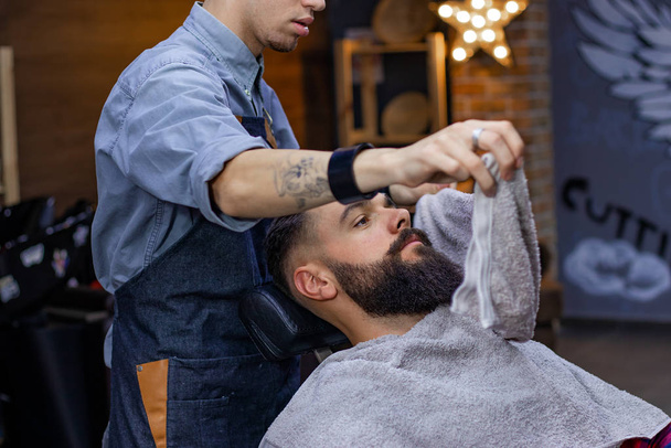 Barbeiro cobre o rosto de um jovem barbudo com uma toalha quente antes de barbear real profissional na barbearia. Conceito de publicidade e barbearia
. - Foto, Imagem