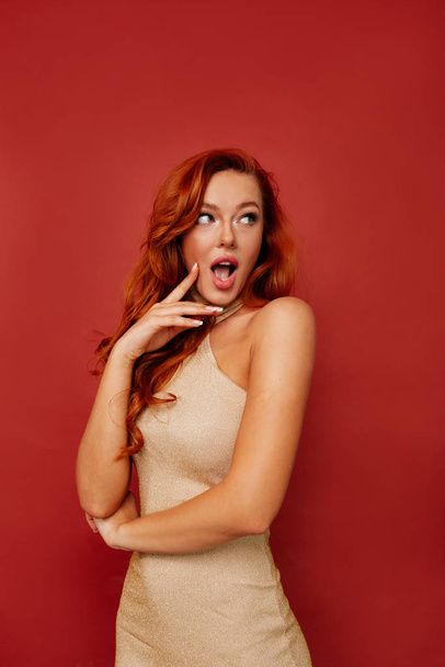 Attractive stylish woman with red hair wearing gold evening dress posing with surprised emotions over red background  - Foto, immagini