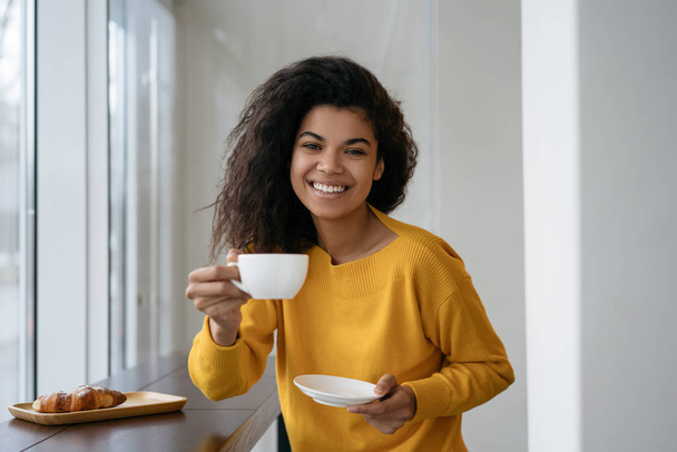 junge attraktive afrikanisch-amerikanische Frau trinkt Kaffee, blickt in die Kamera und lächelt in einem modernen Café. Porträt eines schönen Hipster-Mädchens mit einer Tasse Tee in stilvoller Kleidung  - Foto, Bild