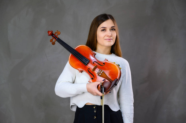 Concept musician violinist. Portrait of a pretty brunette girl in a white sweater playing the violin on a gray background - Foto, imagen