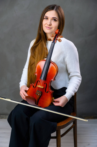 Portrait of a pretty brunette musician girl in a white jacket on a gray background sits on a chair with a violin in her hands - Foto, afbeelding