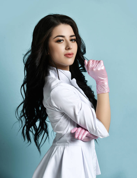 Portrait of sexy brunette woman professional cosmetologist in medical uniform gown is posing sideways holding hand up - Foto, afbeelding
