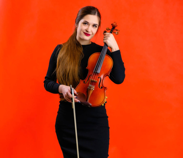 Portrait of a pretty brunette musician girl with a smile in a black dress on a red background holds a violin in her hands - Фото, зображення