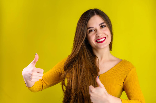 Portrait of a pretty brunette girl with long hair with a smile in a yellow sweater on a yellow background smiles and shows positive emotions - Foto, imagen
