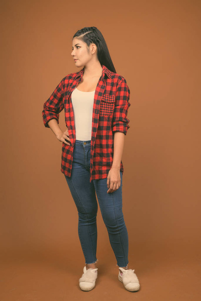 Young beautiful Asian woman wearing red checkered shirt against brown background - Fotó, kép