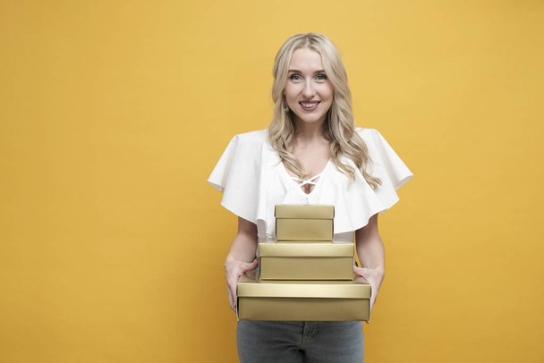 Happy blond european woman in white blouse holding a pack of gift boxes over yellow background. Copy space - Φωτογραφία, εικόνα