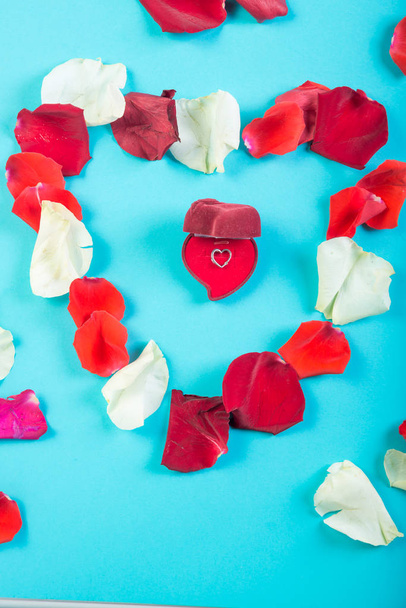 Heart of rose petals on a blue background and a heart-shaped box with a pendant. - Фото, зображення