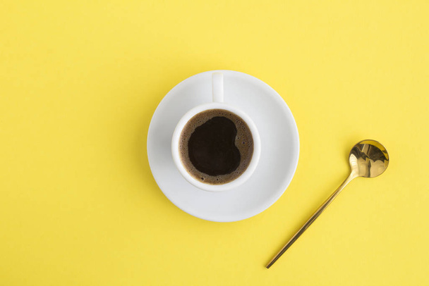schwarzer Kaffee in der weißen Tasse und goldener Löffel in der Mitte des gelben Hintergrunds. Kopierraum. Ansicht von oben. - Foto, Bild