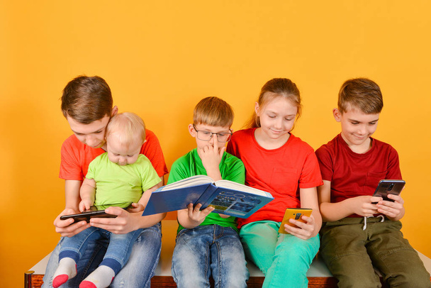 Dzieci ze smartfonem siedzą w pobliżu, jeden chłopiec w okularach siedzi z książką. - Zdjęcie, obraz