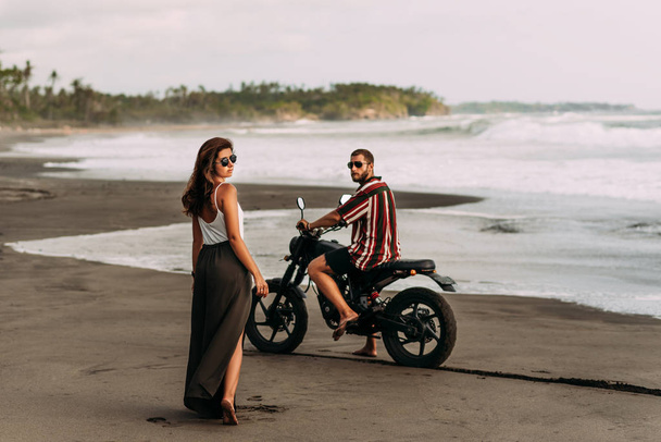 Stylish couple on a motorcycle on the beach. Couple in love at sunset by the sea. Beautiful couple on a motorcycle. Travel on a motorcycle. Lovers at sunset. Journey to the island of Bali - Фото, изображение