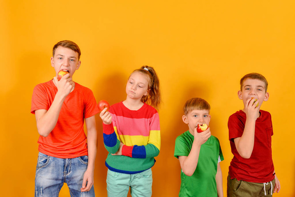 Quatre enfants mangent une pomme debout les uns à côté des autres, les gars sur un fond jaune dans des vêtements de couleur colorée
. - Photo, image
