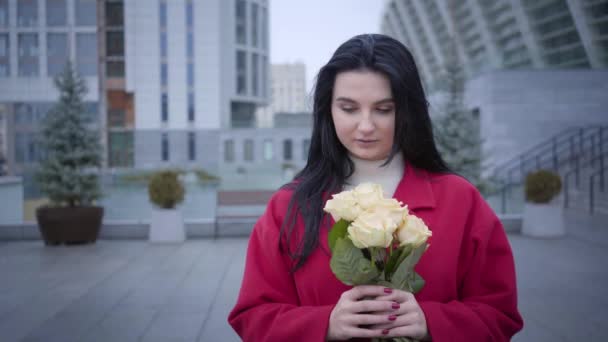 Portrait of romantic young Caucasian woman smelling yellow roses and looking at camera. Fashionable lady with black hair dressed in red coat posing in city in autumn day. Joy, happiness, lifestyle. - Séquence, vidéo