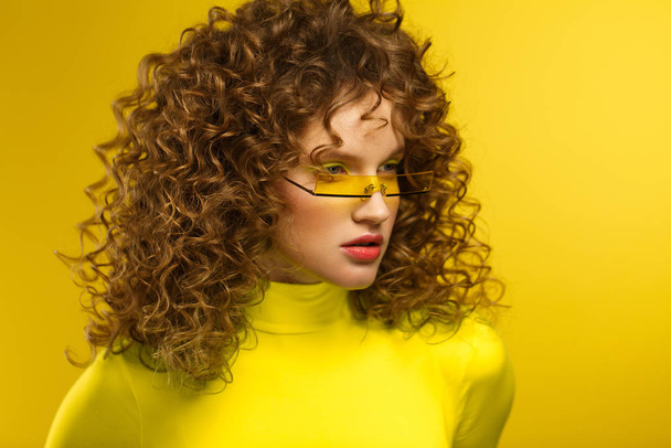 Fashion studio portrait of young beautiful women with curly hair. Nice girl with afro hair and bright make up wearing yellow eyeglasses standing on yellow background. Beauty and hair care concept - Photo, Image